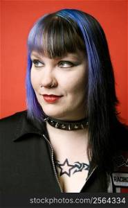 Portrait of Caucasian woman with blue hair, tattoo, and spike collar against orange background.