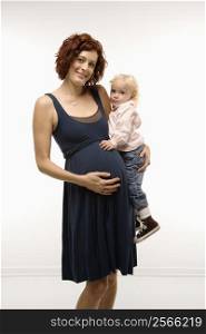 Portrait of Caucasion mid-adult attractive pregnant woman standing, holding female toddler on hip and other hand on belly, looking at viewer and smiling.