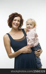 Portrait of Caucasion mid-adult attractive pregnant woman standing, holding female toddler on hip and other hand on belly, looking at viewer and smiling.