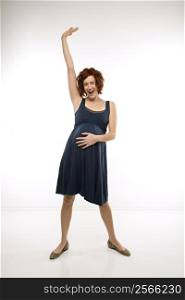 Portrait of Caucasion mid-adult attractive pregnant woman standing with one hand on belly and other arm in air looking at viewer and smiling.