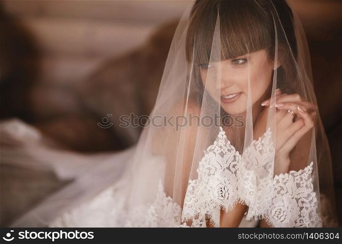 Portrait of charming bride sitting on the bed in a hotel room. the bride is covered with veil. Close up. Wedding morning. Gentle, Tender emotion on the face. Portrait of charming bride sitting on the bed in a hotel room. the bride is covered with veil. Close up. Wedding morning. Gentle, Tender emotion on the face.