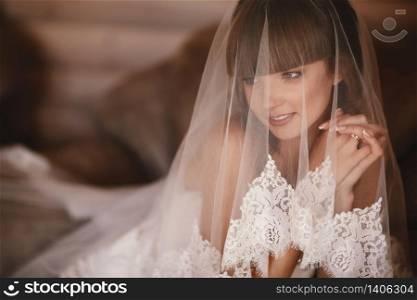 Portrait of charming bride sitting on the bed in a hotel room. the bride is covered with veil. Close up. Wedding morning. Gentle, Tender emotion on the face. Portrait of charming bride sitting on the bed in a hotel room. the bride is covered with veil. Close up. Wedding morning. Gentle, Tender emotion on the face.