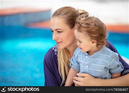 Portrait of charming mother with cute little son sitting near pool, spending summer holidays on the beach resort