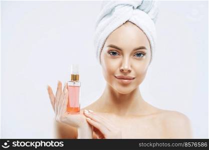 Portrait of charming young healthy woman holds bottle of expensive parfum, enjoys pleasant aroma, has healthy skin, wears bath towel on head, stands topless indoor. Women and cosmetics concept