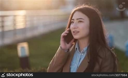 Portrait of cheerful asian girl chatting on smart phone at sunset outdoors. Beautiful hipster young female communicating with her boyfriend on mobile phone while relaxing in the park.