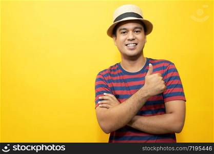 Portrait of cheerful asian man in basic clothing Casual wear red striped T-shirt and wear a hat smiling and show giving thumbs up at camera with showing success. isolated over yellow background.