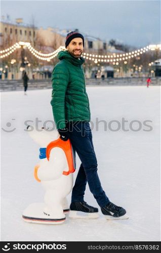 Portrait of cheerful bearded male stands near skate aid, going to skate for first time, being in good mood, enjoys winter holidays. Happy unshaven man in warm clothes and skates stand on ice rink