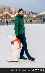 Portrait of cheerful bearded male stands near skate aid, going to skate for first time, being in good mood, enjoys winter holidays. Happy unshaven man in warm clothes and skates stand on ice rink