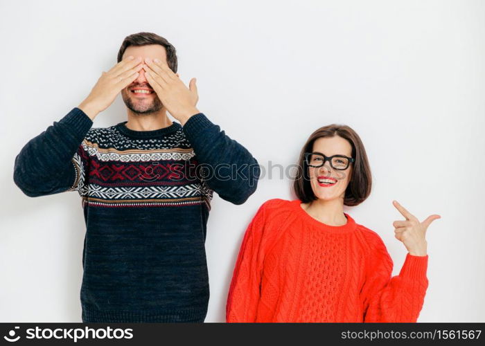 Portrait of cheerful couple pose against white background. Glad brunette female in red sweater indicates at her husband who closes eyes as anticipates for present, Family, relationships, togetherness