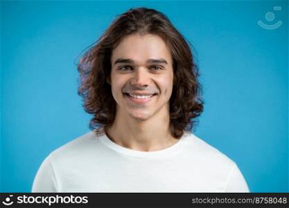 Portrait of cheerful european man looking to camera. Young handsome kind guy with long hairstyle smiling in studio on blue background. High quality photo. Portrait of cheerful european man looking to camera. Young handsome kind guy with long hairstyle smiling in studio on blue background.