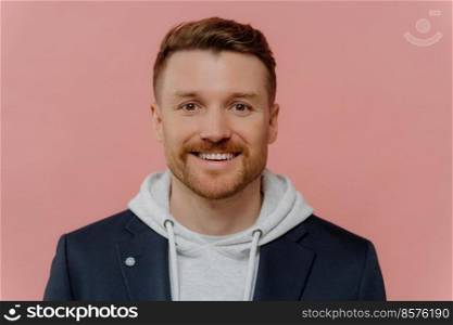 Portrait of cheerful European man with stubble dressed in hoodie and black jacket looks directly at camera has happy face expression poses against pink background. People emotions lifestyle.