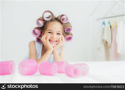 Portrait of cheerful female kid keeps hands under chin, has curlers on hair, going to have nice hairstyle, poses against white background, has charming smile, being in good mood. Children and beauty