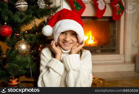 Portrait of cheerful girl in Santa hat sitting by the burning fireplace at house