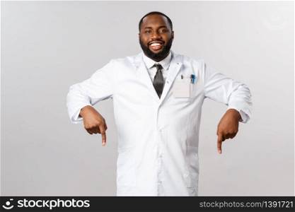 Portrait of cheerful handsome african-american male doctor inviting patients his clinic, check-up or professional treatment, covid19 test, pointing fingers down, smiling give advices during pandemic.. Portrait of cheerful handsome african-american male doctor inviting patients his clinic, check-up or professional treatment, covid19 test, pointing fingers down, smiling give advices during pandemic