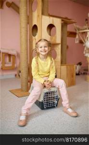 Portrait of cheerful little girl sitting on cage with cat over shelter playground. Child enjoying company of having pet and learning responsibility of animal care. Portrait of cheerful girl sitting on cage with cat over animal shelter