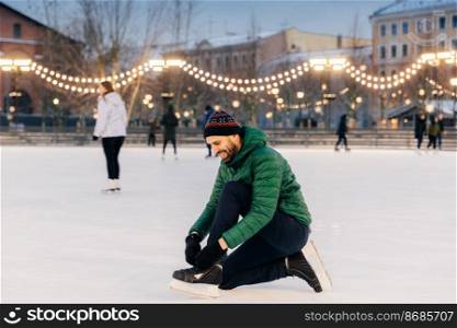 Portrait of cheerful male laces up skates as going to go skating on ice rink, prepares or puts on special shoes, enjoys wonderful winter holidays. Outdoor shot of delightful male skater on ring
