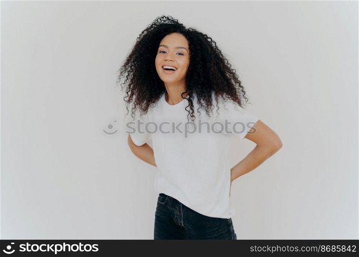 Portrait of cheerful millennial woman keeps hands behind back, dressed in casual wear, laughs from something fun, isolated over white studio background. African American lady expresses happiness