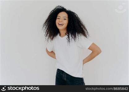 Portrait of cheerful millennial woman keeps hands behind back, dressed in casual wear, laughs from something fun, isolated over white studio background. African American lady expresses happiness