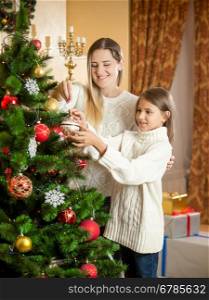 Portrait of cheerful mother with daughter decorating Christmas tree with colorful baubles
