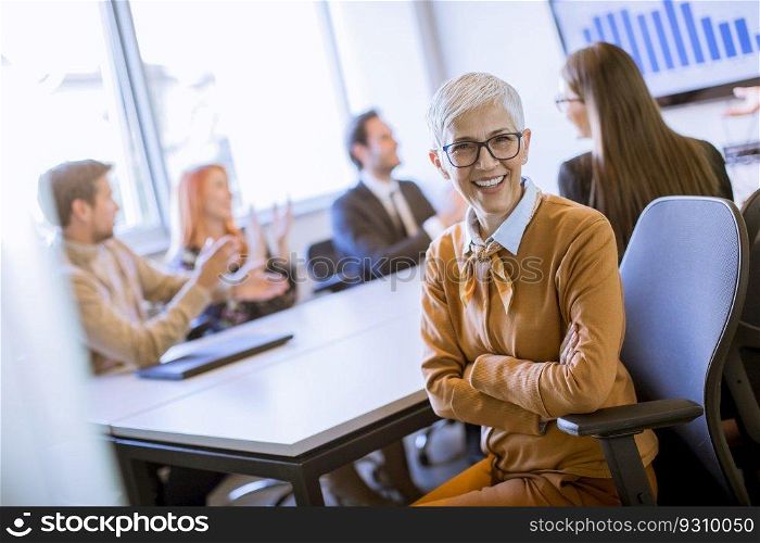 Portrait of cheerful senior businesswoman in office while other business people having meeting