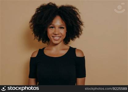 Portrait of cheerful smiling beautiful dark skinned girl in black tshirt standing isolated against dark beige wall background with copy space for advertising. Youth and happiness concept. Portrait of cheerful smiling beautiful dark-skinned girl in black tshirt
