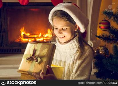 Portrait of cheerful smiling girl sitting by the fireplace and looking inside of present box