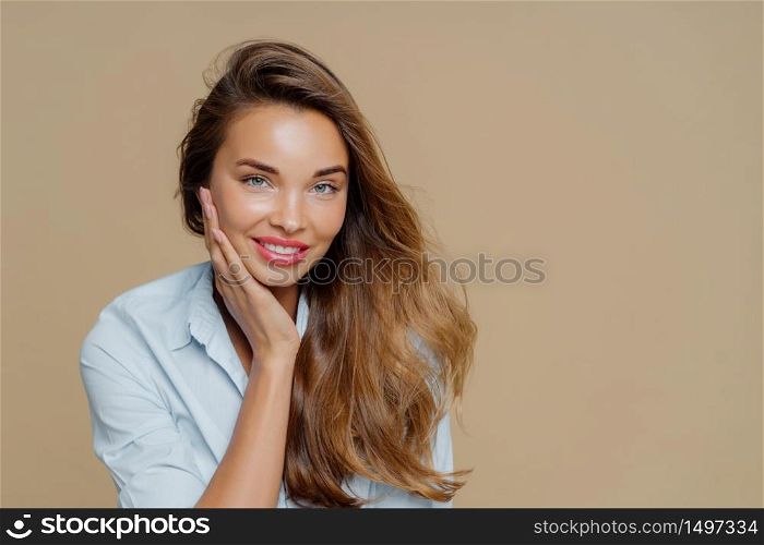 Portrait of cheerful smiling woman touches cheek, has toothy charming smile, wears shirt, looks at camera with pleasure, has long wavy hair, makeup, poses against brown background, free space