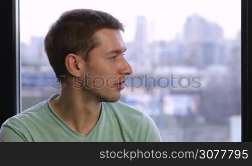 Portrait of cheerful young guy looking out of the window, turning his head and smiling on cityscape background. Positive young hipster with toothy smile looking at the camera at home interior. Positive emotion and facial expression.