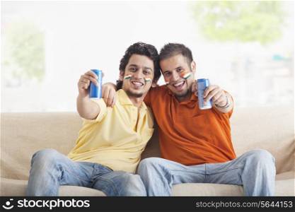 Portrait of cheerful young male friends with face painted holding out tin cans while sitting on sofa
