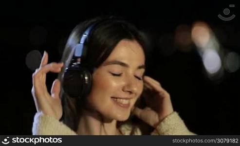 Portrait of cheerful young millennial woman with headphones listening to music outdoors in city night. Joyful beautiful female singing along with the song, enjoying time while listening muisc with earphones over blurry streetlights bokeh background.