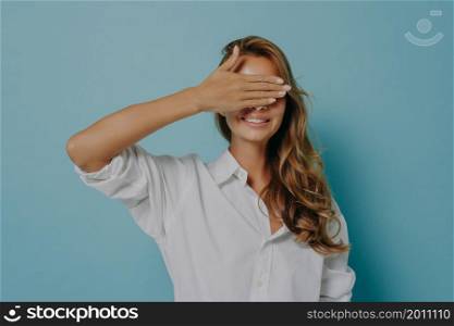 Portrait of cheerful young woman broadly smiling while covering her eyes with hand happily awaiting for welcome surprise or playing hide and seek game, isolated over light blue studio background. Portrait of cheerful young woman broadly smiling while covering her eyes with hand