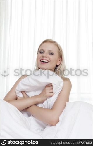 Portrait of cheerful young woman covering herself with bedsheet in bed