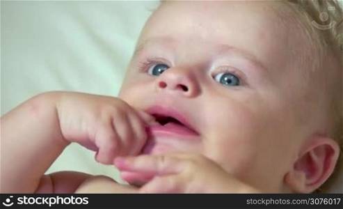 Portrait of child having fun in cradle at home, happy cute little boy laughing and thumb sucking in bed. Male infant with blue eyes in baby crib. Close-up of face, slow motion