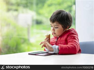 Portrait of child holding ice cream and watching cartoon on mobile phone, Relaxing preschool boy sitting at the table in a cafe with blurry nature background