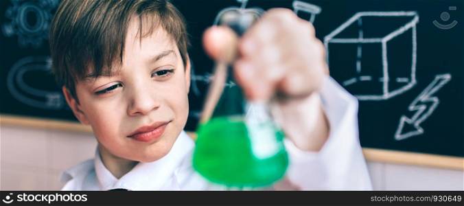 Portrait of concentrated little scientist looking flask with chemical green liquid against of drawn blackboard. Serious boy looking flask with chemical liquid