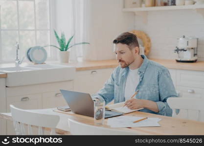 Portrait of concentrated male freelancer working on project remotely from home while sitting at kitchen table with laptop, wears casual clothes. Business online and freelance concept. Portrait of handsome focused businessman working on project remotely from home