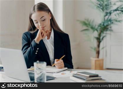 Portrait of concentrated, thinking young caucasian bussiness woman in black formal suit sitting and writing down in note book in front of laptop on white desk with bow-pot in blurred background. Concentrated business woman thinking about work issues at office