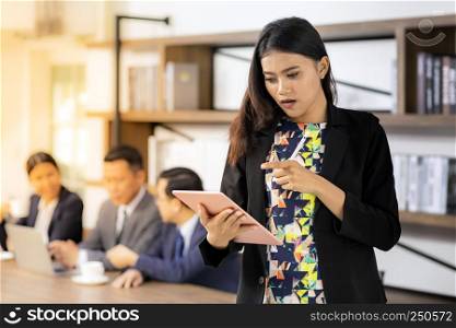 Portrait of confidence asian Businesswoman working and thinking with smart tablet and stand in front of table in meeting room of cafe with business team in background using for coporate background work