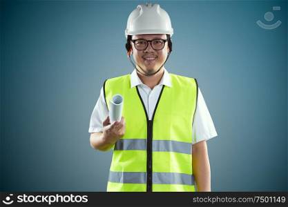 Portrait of confident asian young engineer wearing protective hard hat and holding paper blueprint .