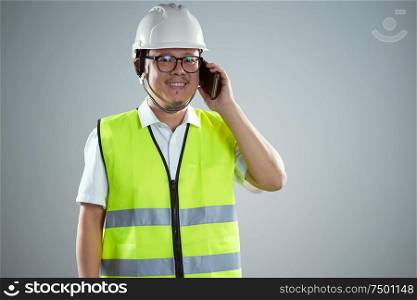 Portrait of confident asian young engineer wearing protective hard hat and using smartphone .