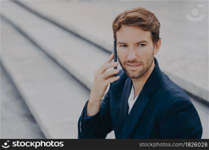 Portrait of confident attractive business proffesional making call, talking on smartphone, having business conversation with partner, looking away while sitting outside on stone stairs in city center. Confident attractive businessman in suit talking on smartphone outside