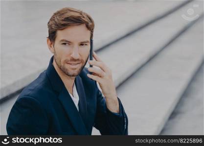 Portrait of confident attractive business proffesional making call, talking on smartphone, having business conversation with partner, looking away while sitting outside on stone stairs in city center. Confident attractive businessman in suit talking on smartphone outside
