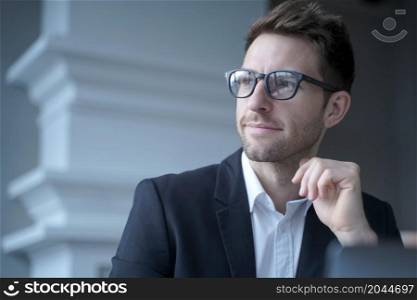 Portrait of confident attractive young european businessman in formal wear sitting at desk in front of laptop at home office, looking aside with pensive facial expression. Business people concept. Confident attractive young businessman sitting at home office, looking aside with pensive expression