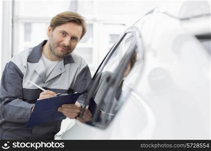 Portrait of confident automobile mechanic holding clipboard while leaning on car&rsquo;s window in workshop