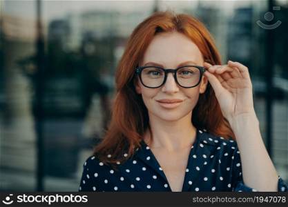 Portrait of confident beautiful red-haired business woman adjusting spectacles eyeglasses while posing on city street in summer, looking at camera with confidence, blurred urban background. Confident beautiful red-haired business woman adjusting spectacles eyeglasses posing on street