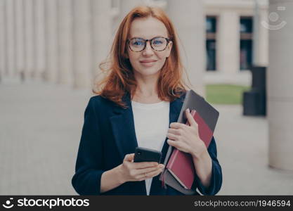 Portrait of confident beautiful red-haired female business consultant holding modern smartphone and laptop, looking at camera with smile, wearing spectacles, standing outdoors after working day. Confident beautiful red-haired female business consultant holding modern smartphone and laptop