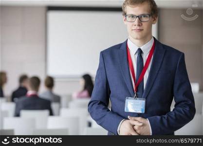 Portrait of confident businessman standing at seminar hall with colleagues sitting in background