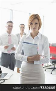 Portrait of confident businesswoman holding book with colleagues in background at office