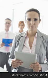 Portrait of confident businesswoman holding digital tablet with colleagues in background at office