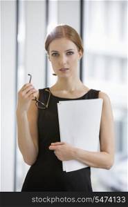 Portrait of confident businesswoman holding documents and eyeglasses in office